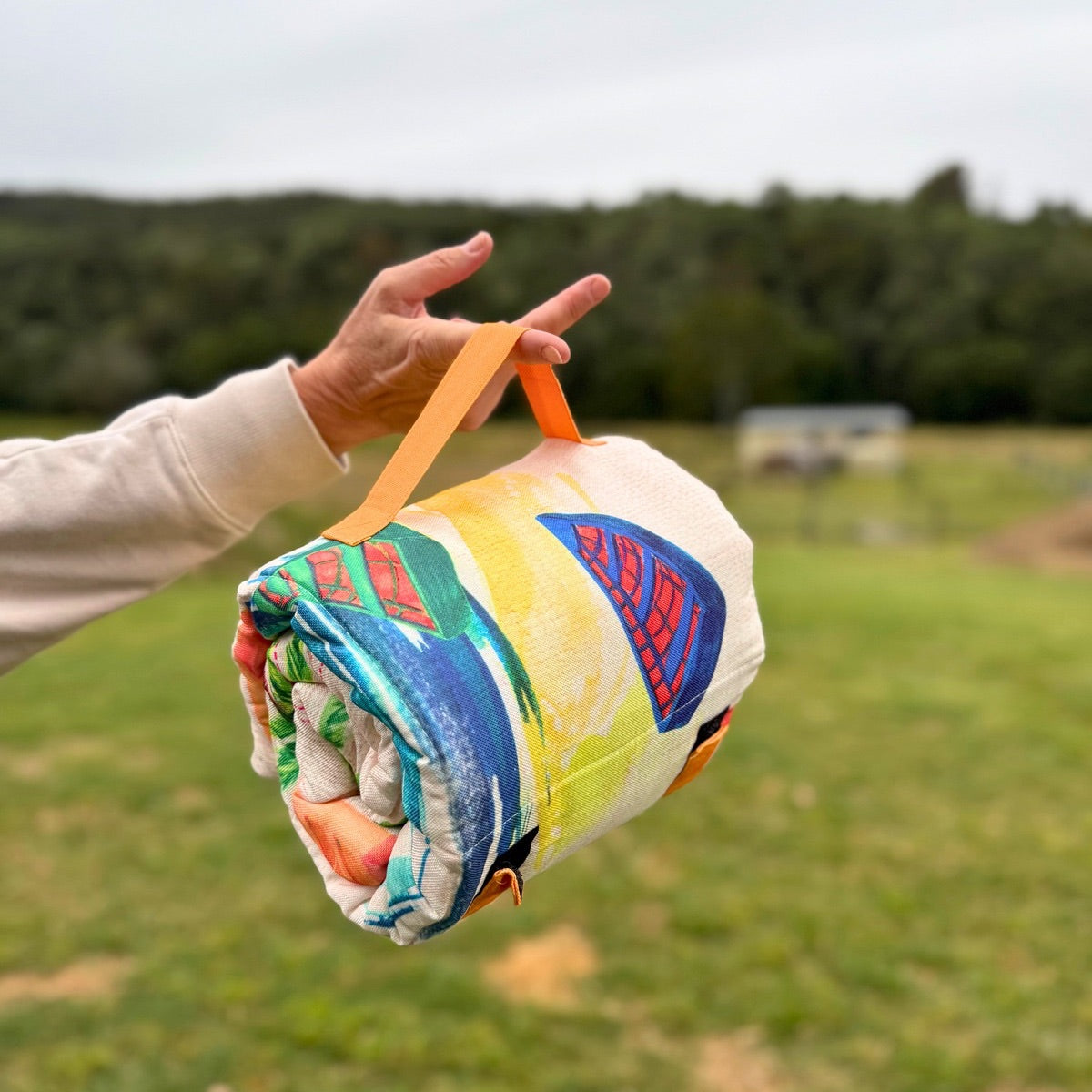 Rolled-up ‘Fire Dog’ training mat, crate liner, cafe bed, and car seat protector, featuring a vibrant tropical-themed design with palm trees, boats, and huts in bright watercolour-style colours, secured with a handle, resting on green grass.