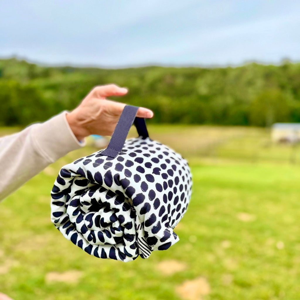 Rolled-up ‘Gigi’ training mat, dog bed, cafe mat, and crate liner, named after a cheeky Cavoodle, featuring a black and white paint spot design with a black handle, held outdoors against a grassy background